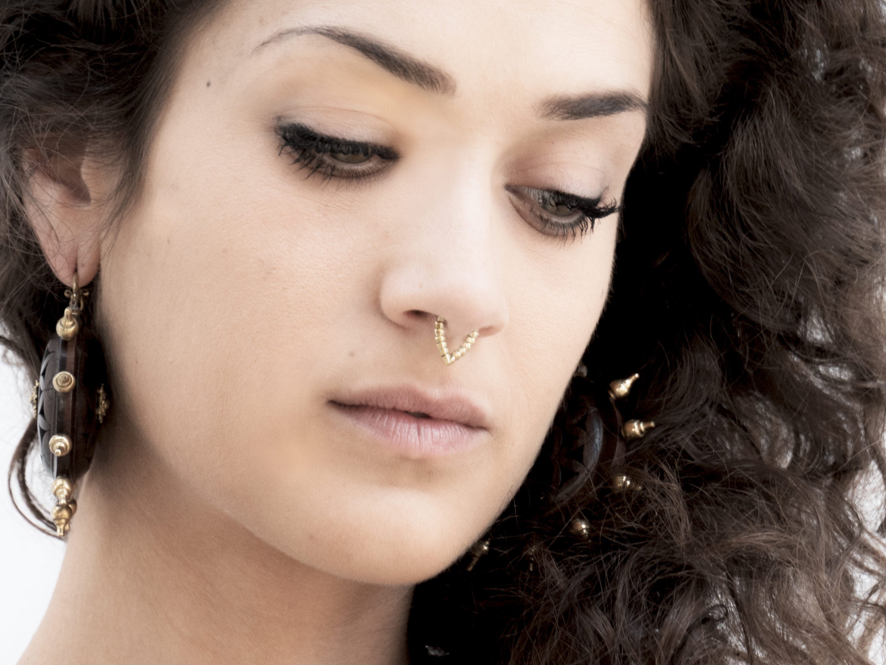 Close-up of a person with curly brown hair looking down, wearing the KAMO Biomech Teardrop Septum Ring in Gold by Kaya Movement and large hoop earrings adorned with beads and spikes. The expression is thoughtful, and the background is neutral.